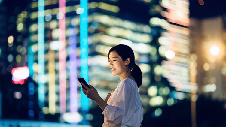 Smiling woman checking mobile phone against glowing city skyscrapers at night